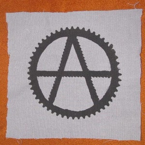 May include: A black and white patch featuring an anarchy symbol within a gear design. The patch is made of fabric and has a grey background.