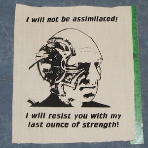May include: Black and white image of a person's head with a mechanical device attached to it. The image is on a beige fabric background. The text "I will not be assimilated! I will resist you with my last ounce of strength!" is printed below the image.