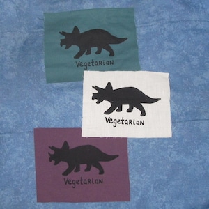 May include: Three fabric squares with a black silhouette of a triceratops dinosaur on each. Each square has the word "Vegetarian" printed below the dinosaur. The squares are green, white, and purple.
