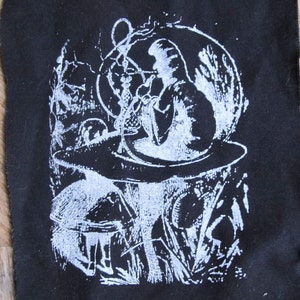 May include: A black and white illustration of the Cheshire Cat from Alice in Wonderland sitting on a mushroom, smoking a hookah. The image is printed on a black fabric patch.