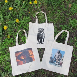 May include: Three white tote bags with different graphic designs. The first bag features a black and white illustration of an owl. The second bag features a black and white illustration of a creature from the black lagoon. The third bag features a color photograph of a woman with a headdress.
