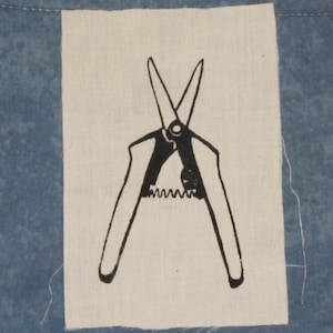 Peut inclure: Illustration en noir et blanc d'un sécateur de jardin sur un patch de tissu crème. Le sécateur est ouvert, avec un mécanisme à ressort visible. Le patch est posé sur un fond bleu, suggérant un article textile ou artisanal.