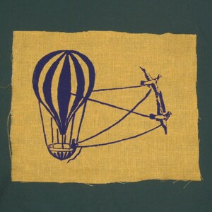May include: A yellow fabric patch with a blue and white hot air balloon design. The balloon is attached to a series of strings that are tied to a branch.