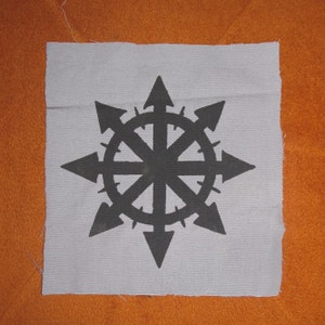 Op de afbeelding: Een zwart-witte patch met een symbool van een cirkel met acht punten, waarbij elk punt een pijlpunt is dat naar buiten wijst. De cirkel is omgeven door een ring van stekels.