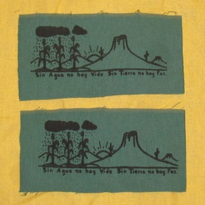Peut inclure: Deux panneaux de tissu vert avec des illustrations noires d'une montagne, du soleil, des nuages de pluie et des arbres. Le texte "Sin Agua no hay Vida. Sin Tierra no hay Paz." est imprimé sur chaque panneau.