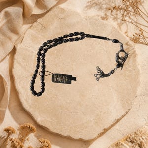 May include: Black and silver prayer beads with a decorative metal tassel. The beads are oval-shaped and strung together with a black cord. A black tag is attached to the beads. The beads are displayed on a beige stone surface.