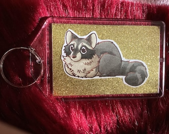 Könnte beinhalten: Ein durchsichtiger Schlüsselanhänger aus Kunststoff mit goldfarbenem Glitzerhintergrund, auf dem ein Cartoon-Waschbär mit schwarzen Augen und einer weißen Brust abgebildet ist.
