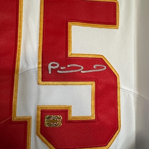 K&ouml;nnte beinhalten: Wei&szlig;es Trikot mit roten und goldenen Akzenten. Die Nummer "15" ist in Rot mit goldenem Rand dargestellt. Eine Unterschrift ist auf dem Trikot zu sehen. Ein goldener Aufkleber ist ebenfalls vorhanden.