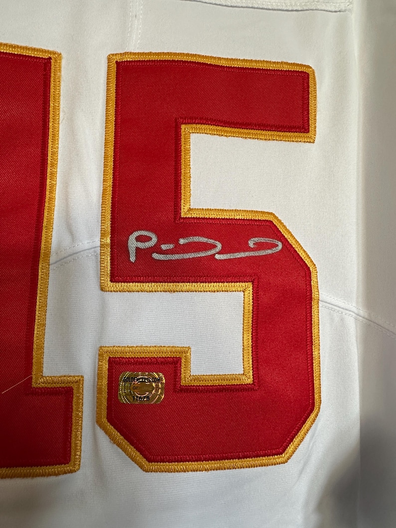 K&ouml;nnte beinhalten: Wei&szlig;es Trikot mit roten und goldenen Akzenten. Die Nummer "15" ist in Rot mit goldenem Rand dargestellt. Eine Unterschrift ist auf dem Trikot zu sehen. Ein goldener Aufkleber ist ebenfalls vorhanden.
