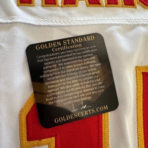 K&ouml;nnte beinhalten: Wei&szlig;es Trikot mit rot-goldenen Stickereien. Das Trikot hat das Wort "MAH" in roten Buchstaben. Eine schwarze Karte mit goldenem Text "GOLDEN STANDARD CERTIFICATION" ist am Trikot befestigt.