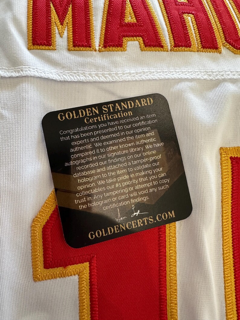 K&ouml;nnte beinhalten: Wei&szlig;es Trikot mit rot-goldenen Stickereien. Das Trikot hat das Wort "MAH" in roten Buchstaben. Eine schwarze Karte mit goldenem Text "GOLDEN STANDARD CERTIFICATION" ist am Trikot befestigt.