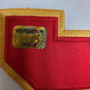 K&ouml;nnte beinhalten: Nahaufnahme eines roten Aufn&auml;hers mit goldfarbenem Rand und einem holografischen Aufkleber. Der Aufkleber lautet "AUTHENTIC GOLDENCERTS.COM GENUINE" und enth&auml;lt die Nummer 11678. Der Aufn&auml;her ist auf einen wei&szlig;en Stoff gen&auml;ht.