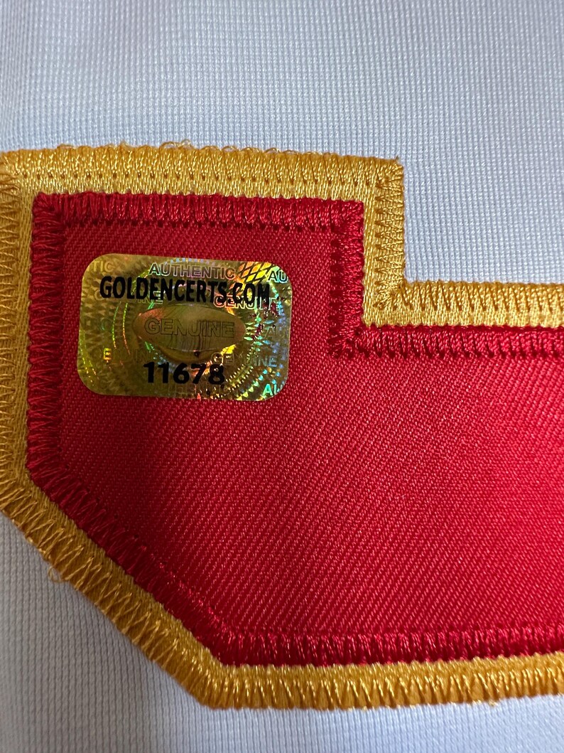 K&ouml;nnte beinhalten: Nahaufnahme eines roten Aufn&auml;hers mit goldfarbenem Rand und einem holografischen Aufkleber. Der Aufkleber lautet "AUTHENTIC GOLDENCERTS.COM GENUINE" und enth&auml;lt die Nummer 11678. Der Aufn&auml;her ist auf einen wei&szlig;en Stoff gen&auml;ht.
