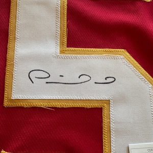 May include: Close-up of a red jersey with a white number patch outlined in gold. The number patch has a black signature. The jersey is made of a breathable fabric.