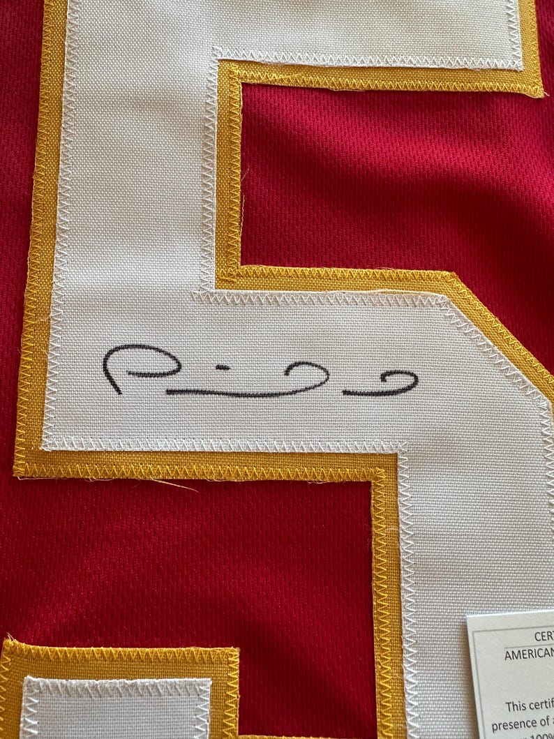 May include: Close-up of a red jersey with a white number patch outlined in gold. The number patch has a black signature. The jersey is made of a breathable fabric.