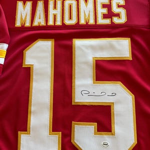 May include: Red football jersey with the name "MAHOMES" in white and gold lettering across the shoulders. The number "15" is prominently displayed in white with gold trim. The jersey is signed in black ink.