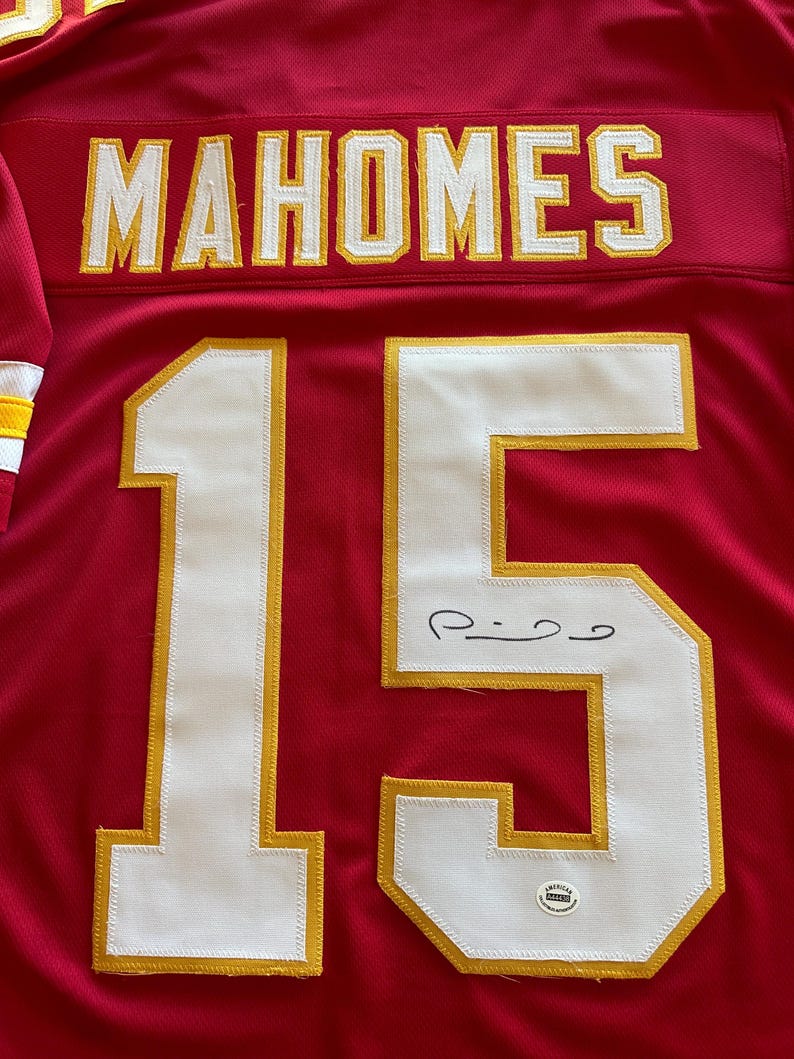 May include: Red football jersey with the name "MAHOMES" in white and gold lettering across the shoulders. The number "15" is prominently displayed in white with gold trim. The jersey is signed in black ink.