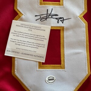 May include: Close-up of a red jersey with a large white number eight patch outlined in gold. The patch has a stitched border and a signature in black ink. A certificate of authenticity is attached, along with a round sticker.