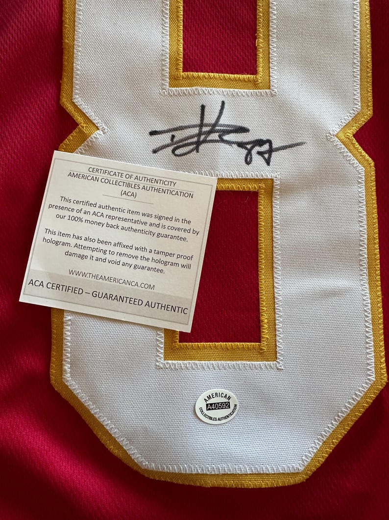May include: Close-up of a red jersey with a large white number eight patch outlined in gold. The patch has a stitched border and a signature in black ink. A certificate of authenticity is attached, along with a round sticker.