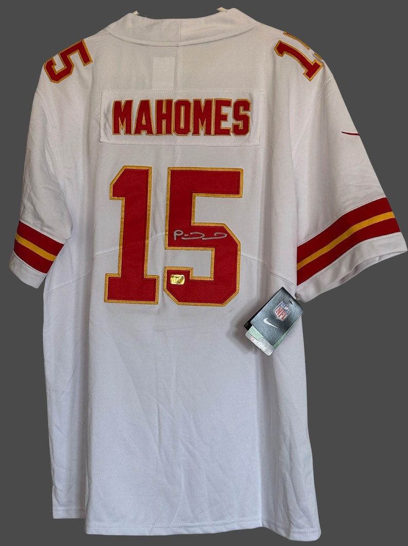 K&ouml;nnte beinhalten: Wei&szlig;es Football-Trikot mit roten und goldenen Akzenten. Das Trikot zeigt den Namen "MAHOMES" und die Nummer "15" auf dem R&uuml;cken sowie eine Unterschrift. Die &Auml;rmel haben rot-goldene Streifen. Ein Nike-Etikett ist angebracht.