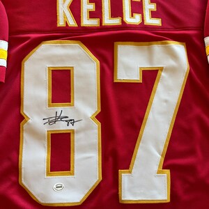 May include: A red jersey with the name "KELCE" in yellow lettering across the shoulders. The number "87" is prominently displayed in white with yellow trim. A signature is visible above the number, and a small authentication sticker is present.