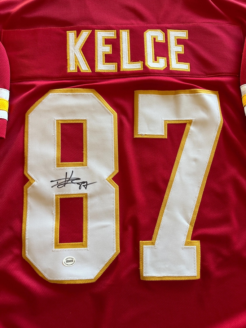 May include: A red jersey with the name "KELCE" in yellow lettering across the shoulders. The number "87" is prominently displayed in white with yellow trim. A signature is visible above the number, and a small authentication sticker is present.
