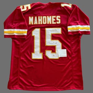 May include: A red football jersey with the name "MAHOMES" and the number "15" in white and gold. The jersey has a signature and a small tag attached. The sleeves have gold and white stripes.