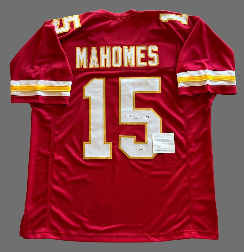 May include: A red football jersey with the name "MAHOMES" and the number "15" in white and gold. The jersey has a signature and a small tag attached. The sleeves have gold and white stripes.