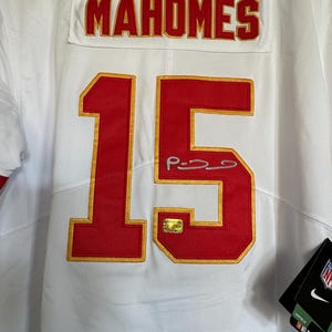 K&ouml;nnte beinhalten: Wei&szlig;es Trikot mit dem Namen "MAHOMES" in roter Schrift auf dem R&uuml;cken und der Nummer "15" in Rot mit goldenem Rand. Das Trikot hat ein Autogramm und einen goldenen Aufkleber. Ein schwarzes Etikett mit den Logos von Nike und NFL Players ist angebracht.
