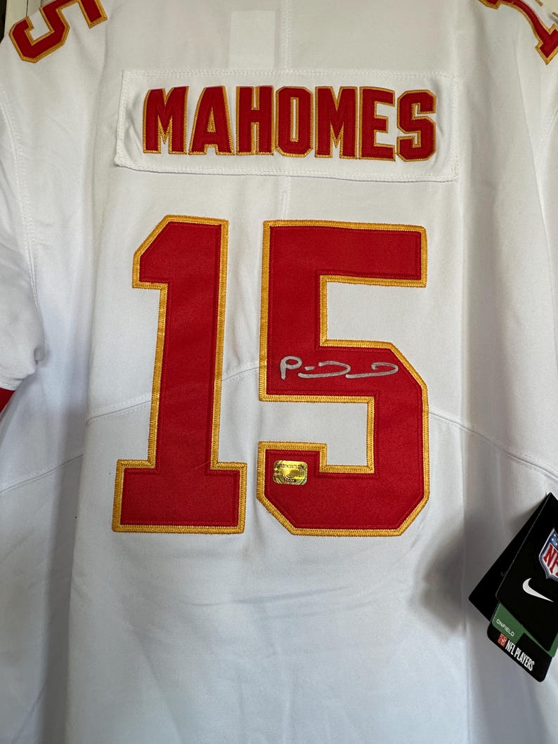 K&ouml;nnte beinhalten: Wei&szlig;es Trikot mit dem Namen "MAHOMES" in roter Schrift auf dem R&uuml;cken und der Nummer "15" in Rot mit goldenem Rand. Das Trikot hat ein Autogramm und einen goldenen Aufkleber. Ein schwarzes Etikett mit den Logos von Nike und NFL Players ist angebracht.