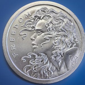 Peut inclure: Une pièce circulaire de couleur argentée présentant le profil en relief d'une personne aux longs cheveux ondulés. Le mot FREEDOM est gravé le long du bord gauche. Un petit symbole triquetra complexe est visible sur l'épaule de la figure.