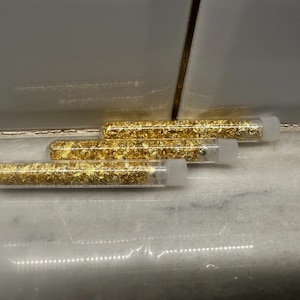 May include: Three clear plastic tubes filled with gold leaf flakes. The tubes are arranged in a staggered row, with white caps on the ends. The gold leaf inside the tubes appears to be a bright, metallic gold color. The tubes are resting on a white surface.