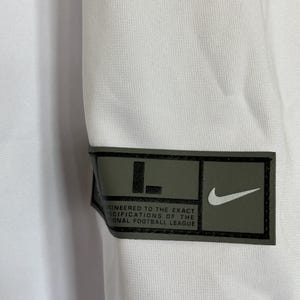 K&ouml;nnte beinhalten: Wei&szlig;es Sporttrikot mit einem schwarzen und olivgr&uuml;nen Aufn&auml;her. Der Aufn&auml;her zeigt den Buchstaben "L", einen Nike-Swoosh und den Text "ENGINEERED TO THE EXACT SPECIFICATIONS OF THE NATIONAL FOOTBALL LEAGUE."