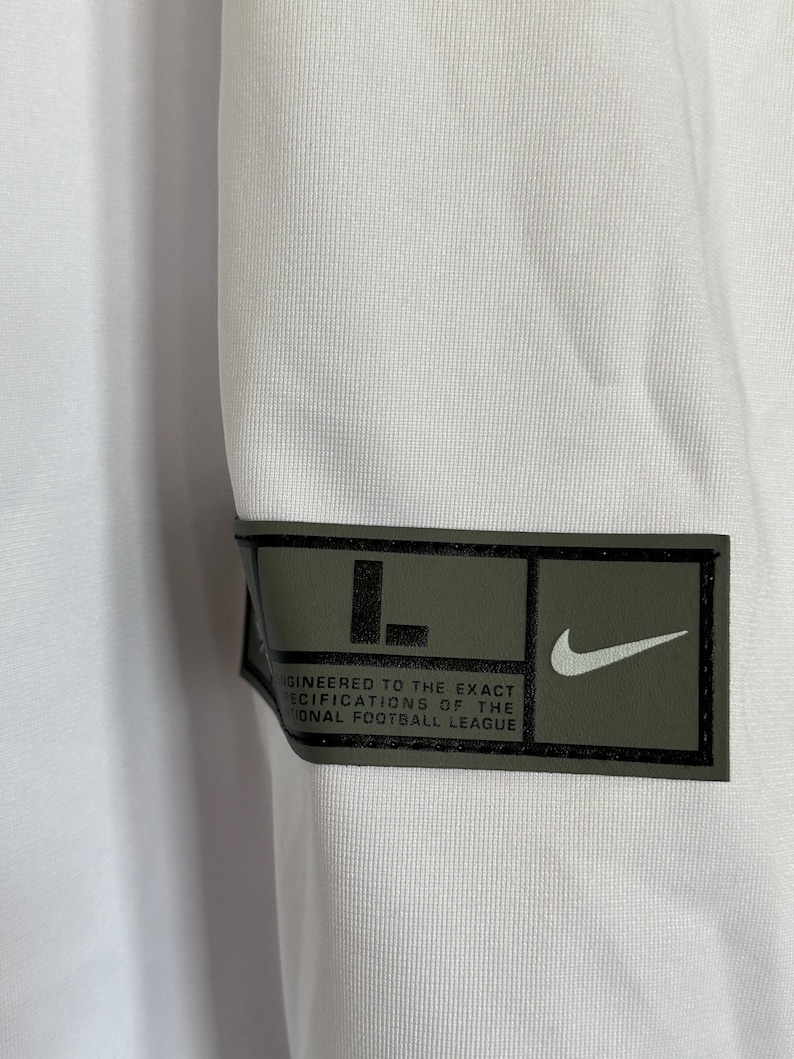 K&ouml;nnte beinhalten: Wei&szlig;es Sporttrikot mit einem schwarzen und olivgr&uuml;nen Aufn&auml;her. Der Aufn&auml;her zeigt den Buchstaben "L", einen Nike-Swoosh und den Text "ENGINEERED TO THE EXACT SPECIFICATIONS OF THE NATIONAL FOOTBALL LEAGUE."