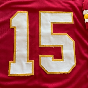 May include: Red football jersey with the number 15 in white with gold trim. The jersey has a V-neck and a mesh-like texture. The sleeve has white, yellow, and red stripes.