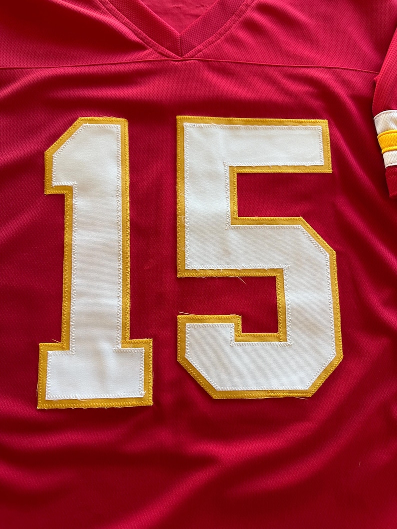 May include: Red football jersey with the number 15 in white with gold trim. The jersey has a V-neck and a mesh-like texture. The sleeve has white, yellow, and red stripes.
