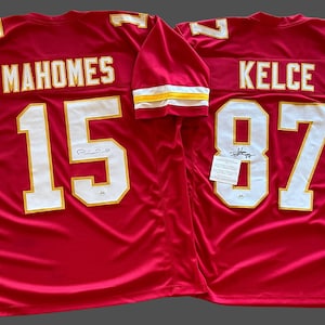 May include: Two red football jerseys. One jersey has "MAHOMES" and the number "15" in white with gold trim. The other jersey has "KELCE" and the number "87" in white with gold trim. Both jerseys have white and gold trim on the sleeves.