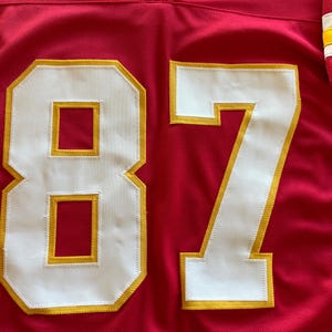 May include: A red football jersey with the number 87 in white with gold trim. The jersey has a V-neck collar and a textured fabric. The number is large and prominently displayed on the front of the jersey.