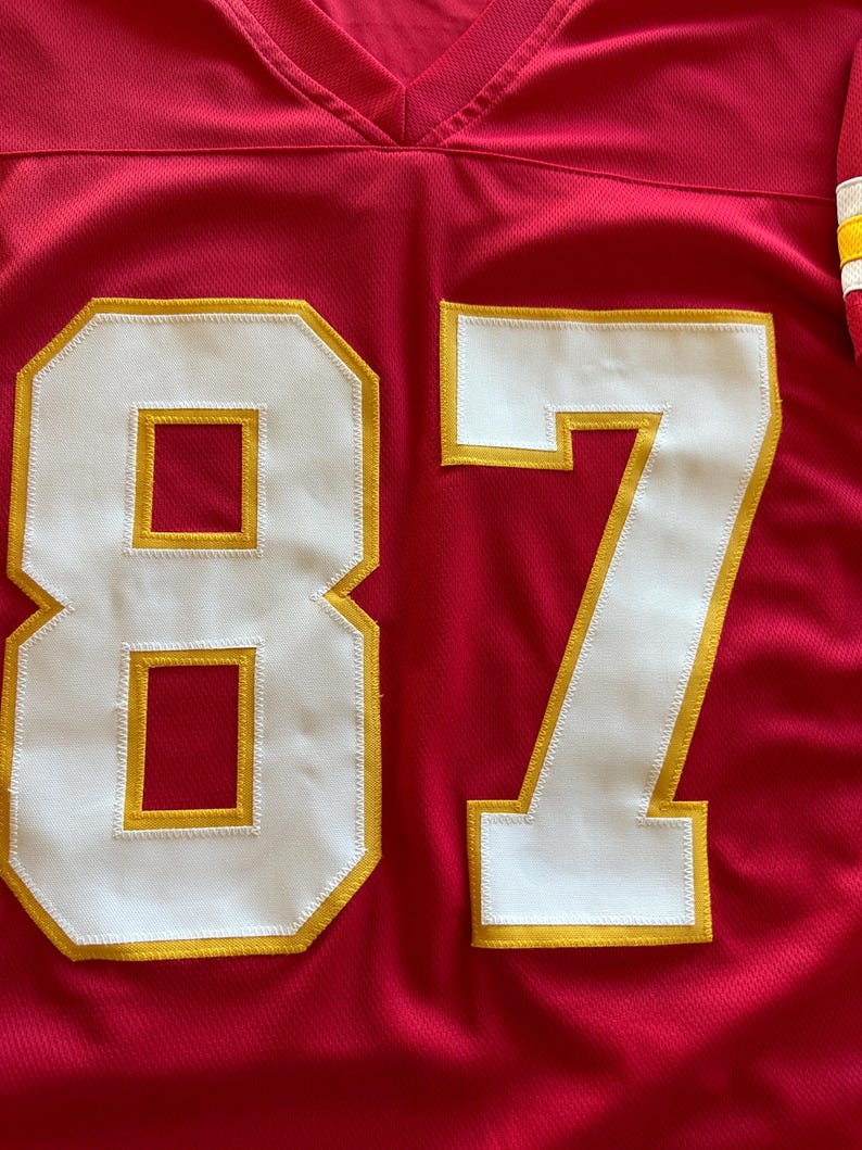 May include: A red football jersey with the number 87 in white with gold trim. The jersey has a V-neck collar and a textured fabric. The number is large and prominently displayed on the front of the jersey.