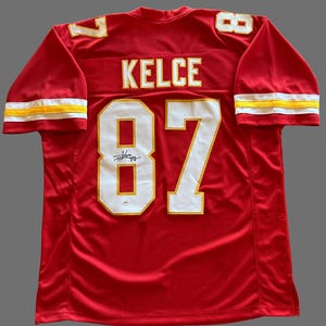 May include: Red football jersey with white and gold accents. The jersey features the name "KELCE" and the number "87" on the back. The sleeves have white and gold stripes. A signature is visible below the number.