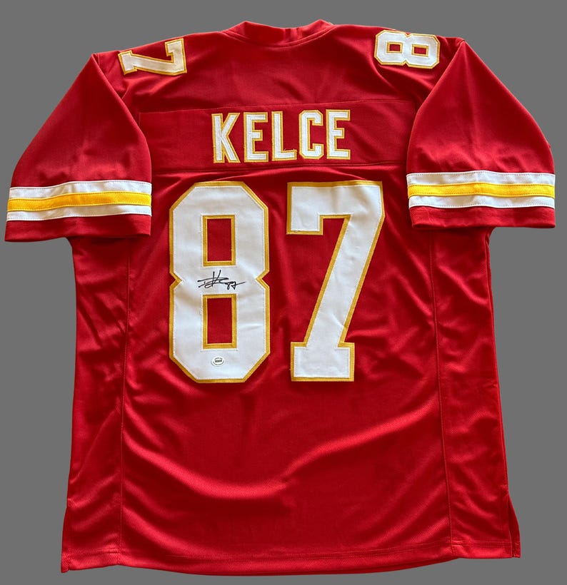 May include: Red football jersey with white and gold accents. The jersey features the name "KELCE" and the number "87" on the back. The sleeves have white and gold stripes. A signature is visible below the number.