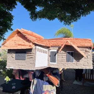 May include: A handmade house costume with a brown shingled roof and grey siding. The costume features a white garage door, windows, and a door. The house is being worn by a person, with a blue sky in the background.