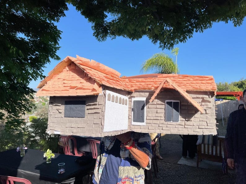 May include: A handmade house costume with a brown shingled roof and grey siding. The costume features a white garage door, windows, and a door. The house is being worn by a person, with a blue sky in the background.