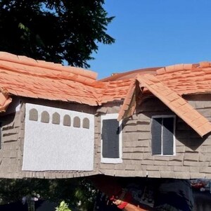 May include: A large, handmade house-shaped pi&ntilde;ata with a brown shingle exterior and a reddish-orange roof. The pi&ntilde;ata features a white garage door, black windows, and a gable roof design. The pi&ntilde;ata is outdoors, with a person in the background.