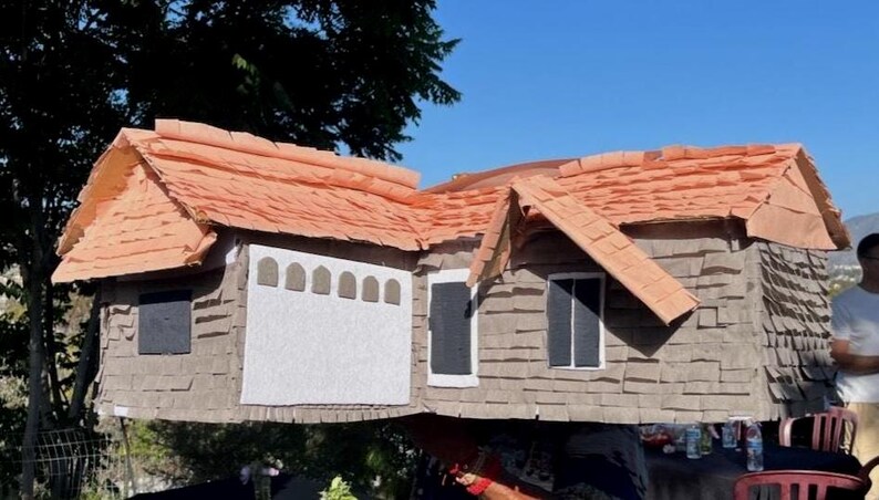 May include: A large, handmade house-shaped pi&ntilde;ata with a brown shingle exterior and a reddish-orange roof. The pi&ntilde;ata features a white garage door, black windows, and a gable roof design. The pi&ntilde;ata is outdoors, with a person in the background.
