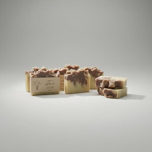 May include: Several bars of handmade soap with a light brown and cream color scheme. Some bars have a label with text and a botanical illustration. The soaps are arranged on a white surface.