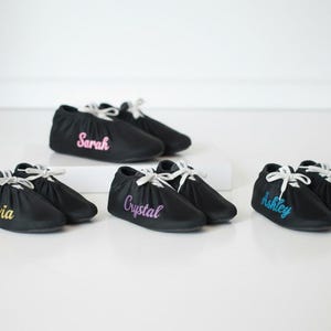May include: Black baby moccasins with white laces. Each pair has a name embroidered in a different color: Olivia (gold), Sarah (pink), Crystal (purple), and Ashley (blue).