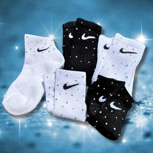May include: An assortment of white and black ankle socks, each adorned with a small black swoosh logo and sparkling embellishments. The socks are arranged against a blue background with star-like light effects.