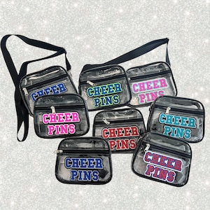 May include: Transparent crossbody bags with black straps and trim. Each bag displays the text "CHEER PINS" in different colours, such as blue, green, pink, and red. The bags have a zip closure and are designed for carrying small items.