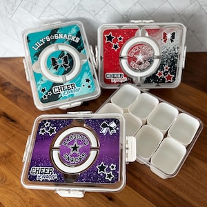 May include: Four clear plastic snack containers with lids. Each lid features a different design with the words "Lily's Snacks", "Kennedy's Snacks", and "Kassidy's Snacks". The containers have a cheerleading theme with stars and bows.